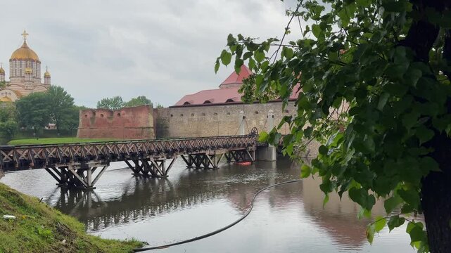 Leafy foreground framing the wooden bridge and brick bastions of Fagaras Fortress reflected in the moat, with a golden Orthodox church dome in the distance.