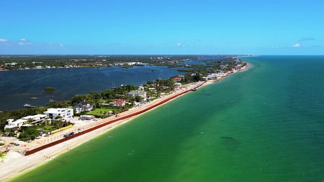 A narrow strip of land lined with houses, a road and a long seawall separates a dark blue bay from green open water.