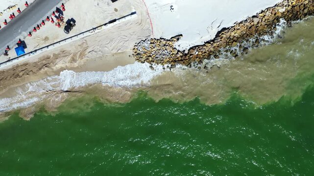 Waves wash over a sandy shoreline beside a rock revetment, with a road, vehicles and traffic cones visible behind the beach.