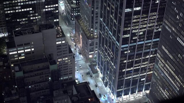 An elevated night view looks between Manhattan office towers toward a brightly lit street in New York City. Rows of windows form geometric patterns in the dark cityscape.