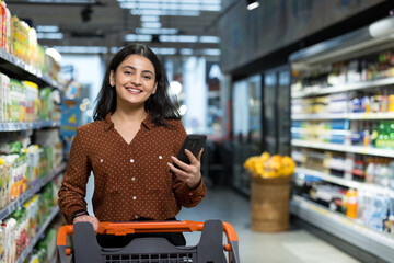 Naklejka na ściany i meble Young indian woman smiling while pushing a shopping cart and using a smartphone to browse products and compare prices in a modern grocery store aisle, candid and cheerful