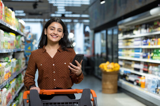 Young indian woman smiling while pushing a shopping cart and using a smartphone to browse products and compare prices in a modern grocery store aisle, candid and cheerful