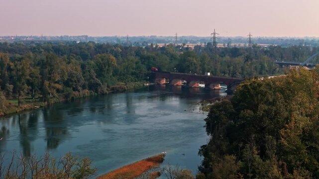 Panoramic aerial view of the adda river flowing through a lush forest, with a truck crossing a historic brick bridge in the distance