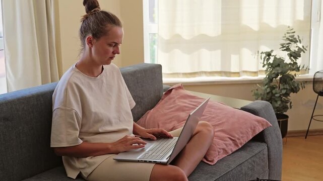 A woman sits on a sofa in her living room, typing on a laptop. Sunlight comes through the window. A plant is in the background near the window
