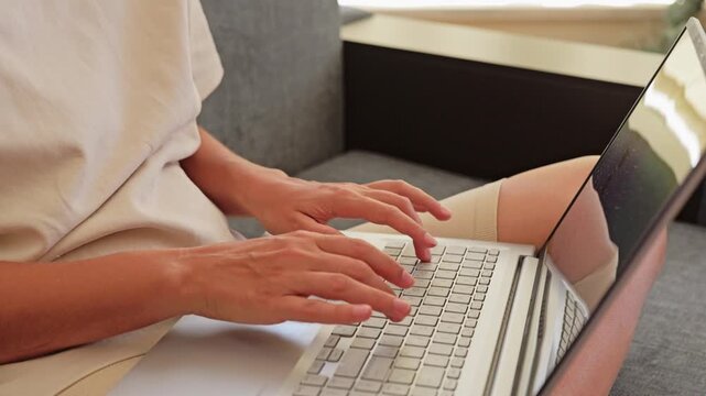 A person sits on a couch and works on a laptop. The sunlight enters the room, creating a bright atmosphere. Hands are positioned on the keyboard