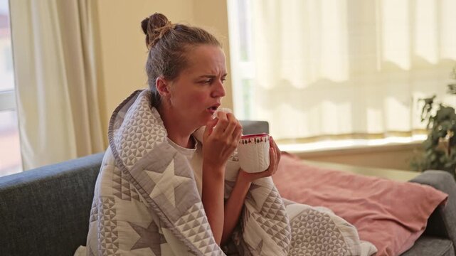 A woman sits on a couch in a living room, holding a mug. She is wrapped in a blanket. Sunlight comes through the window and she appears to be thinking