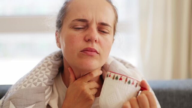 A woman is sitting on a couch at home under a blanket, holding a mug and feeling her throat. She looks uncomfortable and appears to be unwell