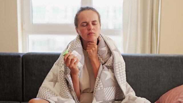 A woman sits on a sofa covered in a blanket. She uses a nasal spray while looking focused. Sunlight comes through a window nearby