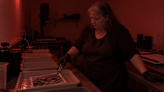 A woman wearing a dark apron leans over a table surface, carefully placing small rounded stones onto a shallow tray using tongs, the entire scene lit with deep red light