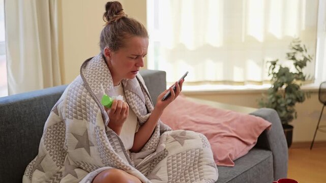 A woman sits on a gray couch covered with a quilt. She holds a green bottle and looks at her tablet. Soft light comes through the window