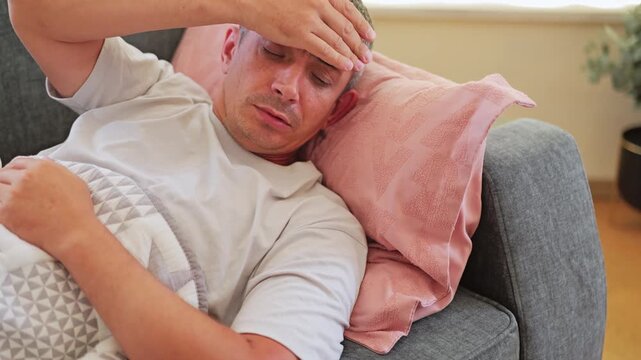 A man is lying on a couch in a living room, looking uncomfortable with his hand on his forehead. He appears to be feeling sick and tired