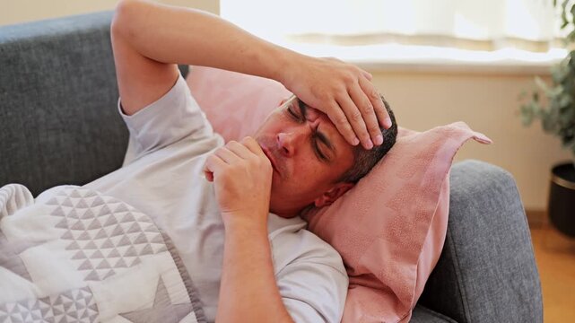 A man rests on a sofa with a headache in his living room. He covers himself with a quilt and keeps his hand on his forehead. The room has natural light and plants