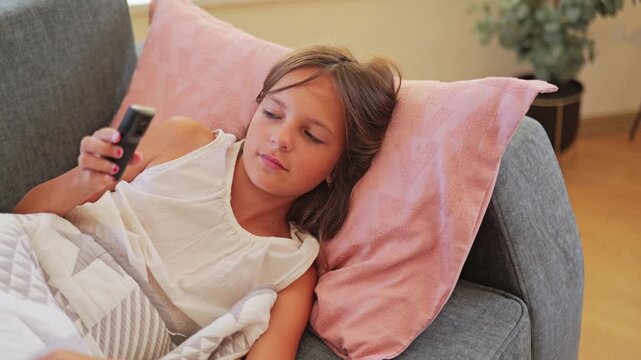 A girl lies on a couch covered with a blanket. She holds a thermometer to her forehead. The room has simple decor and soft colors