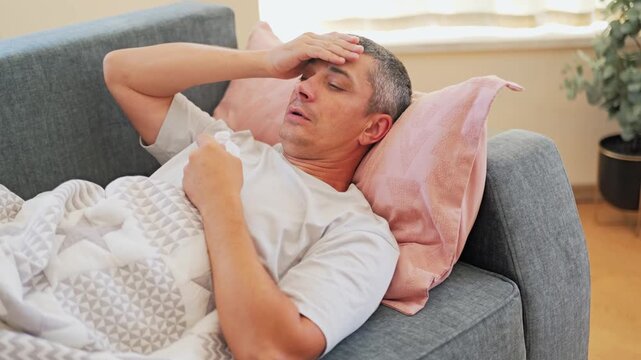 A man is lying on a couch with a blanket, holding his forehead and looking uncomfortable. It is daytime, and he is in a living room with soft light