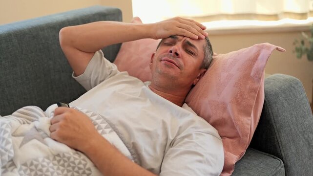 A man is resting on a gray sofa at home. He has a tired expression while yawning. The space has soft pillows and some indoor plants