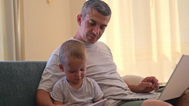 A father is sitting on a couch in a living room. His young child is next to him, looking at some papers. The room is bright with natural light