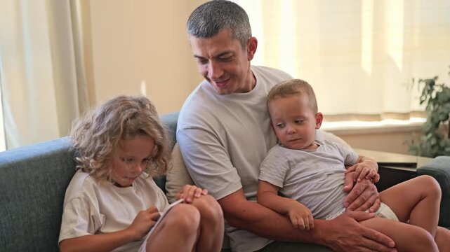 A father sits on a couch with his two children in a well-lit room. The children focus on their activities while the father watches them