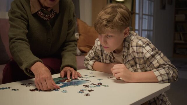 Caucasian senior woman and teenager assembling jigsaw puzzle at table at home. Footage is suitable for family bonding, cognitive health and intergenerational connection campaigns