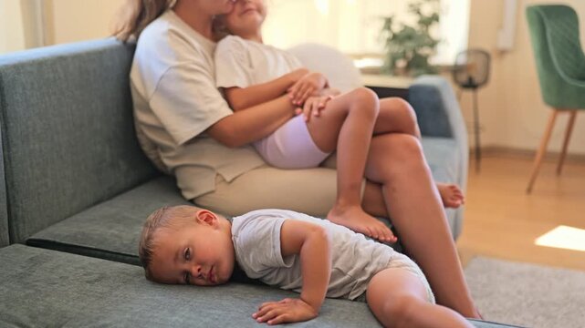 A woman sits on a couch with two children. One child sits on her lap while the other lies on the floor. Sunlight passes through the window