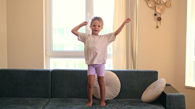 A child stands on a couch and dances joyfully in a room filled with light. The windows show a green view and the decor is simple and modern