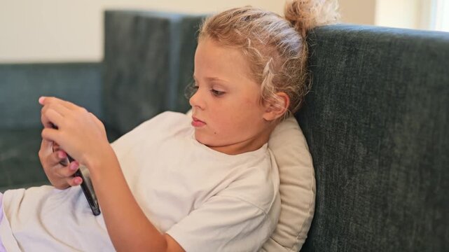 A child is lying on a couch in a living room while looking at a smartphone. The child appears focused on the screen