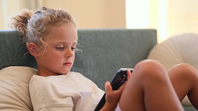 A child reads on her smartphone while sitting on a couch. It is a sunny afternoon with light coming through the window