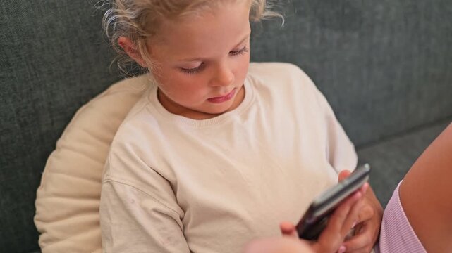 A child sits on a couch, looking closely at a smartphone. The setting is indoors with natural light coming in. The child appears engaged in the device