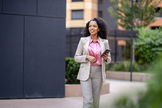 Smiling professional woman walking with a smartphone through a modern business district, representing commuting, mobile work, career confidence and urban professional life