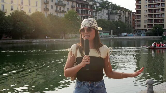 Young female reporter with a microphone passionately speaking to the camera, broadcasting a live news report or a travel vlog from the scenic darsena canal in milan, italy during a sunny day