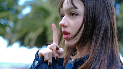 Young woman raising her index finger with a serious expression, side profile portrait outdoors, attention gesture, warning, idea, remember and wait a moment concept, long dark hair, blurred green back © Ethan Blake Archive