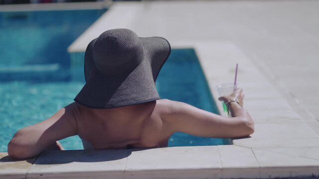 A woman is sitting in a pool with a straw in a green mojito cocktail drink. She is wearing a straw hat and a bikini, cinematic isolated shot