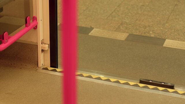 View of commuter train interior door threshold with sliding door partially open to platform, yellow and black safety strip along step, plus pink grab pole and handrail visible.