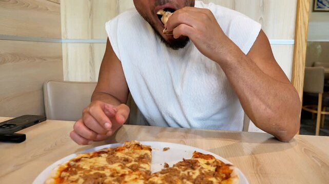 Hungry Young Athletic Man Biting into Hot Slice of Fresh Baked Tuna Pizza at Italian Restaurant Table Fast Casual Food Concept