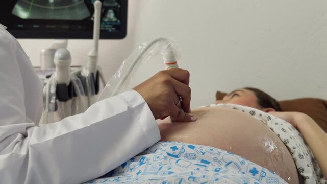 A medical specialist performs a routine sonogram on a 9-month pregnant young Latina woman lying down in a clinic. Both are calm while looking at the screen.