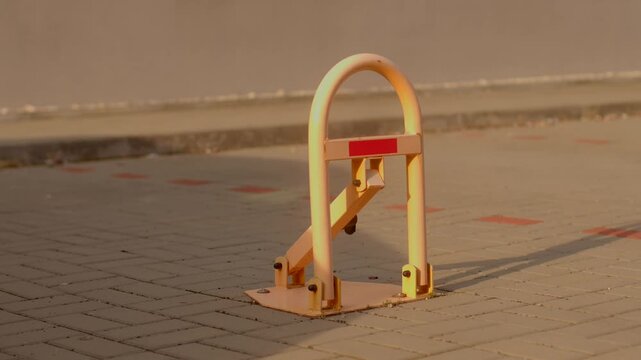 Sunlit yellow metal parking bollard stands locked upright on brick driveway with visible bolts and reflective strip along barrier.
