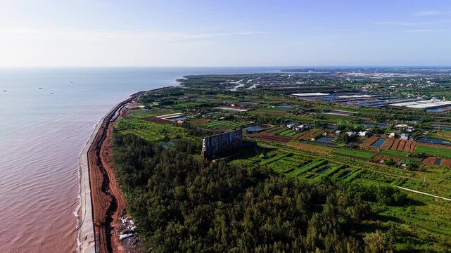 People walk along the shore at Thua Duc Beach. The land features green fields and trees. The sky is clear and blue, with some clouds present. This location shows daily life in the area.