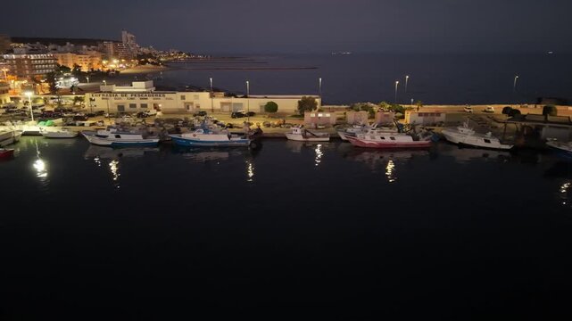 Aerial drone shot moving parallel along the illuminated fishing dock of Santa Pola at night. Features parked boats, glowing harbor lights, and calm waters in Alicante, Spain.