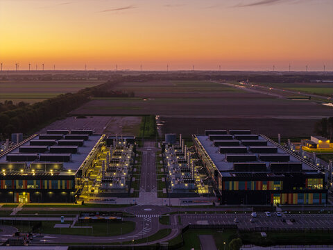 Aerial view of the Microsoft datacenter at sunset, featuring illuminated industrial buildings, vast fields, and wind turbines in Middenmeer, Noord-Holland, Netherlands.