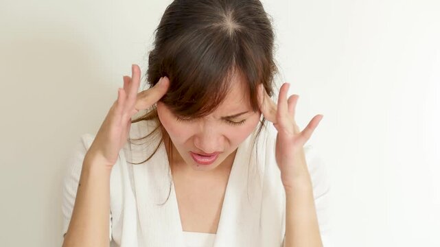 asian woman in her 30s suffering from headache and migraine indoors, touching head with tired and painful expression