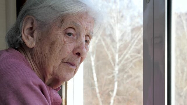 Old woman with deep wrinkles and thoughtful expression is looking out window at street. Elder female face filled with contemplation and loneliness, reflecting on past memories and life experiences