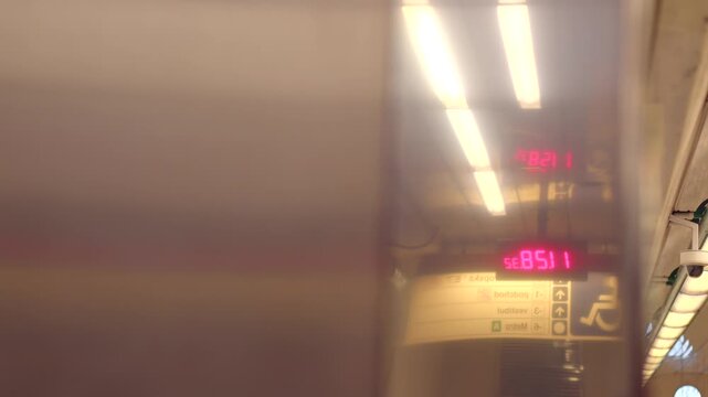 Reflective metal panel inside subway station with warm overhead lights, red digital clock, wayfinding signage, and visible platform edge.