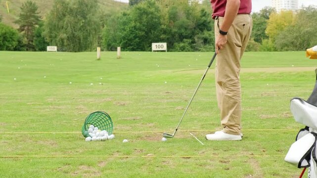 man training on driving range lawn takes careful stance over white golf ball preparing disciplined iron swing beside practice basket full of golf balls and club bag during sunny daytime sport
