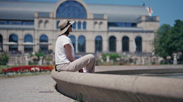 A woman is sitting on a stone edge of Botanical garden fountain in Cologne in front of a large building. She is wearing a hat and a white shirt, cinematic isolated slow motion shot