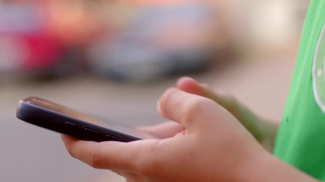 Close-up view of student hands tapping smartphone outside on street in daylight, with blurred background and shallow depth of field.