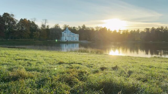 Tracking shot of the Grotto Pavilion across the calm pond on gimbal. Smooth sideways camera movement reveals a serene, sunlit landscape. Located in Catherine Park, Pushkin, Saint Petersburg.