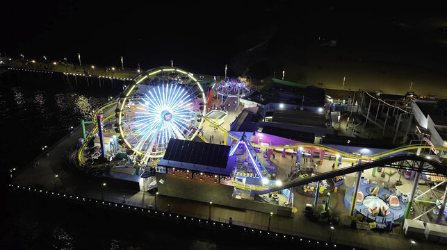 Aerial view of the illuminated Santa Monica Pier at night featuring the glowing Ferris wheel and amusement park rides on the wooden boardwalk in Santa Monica, CA, United States.