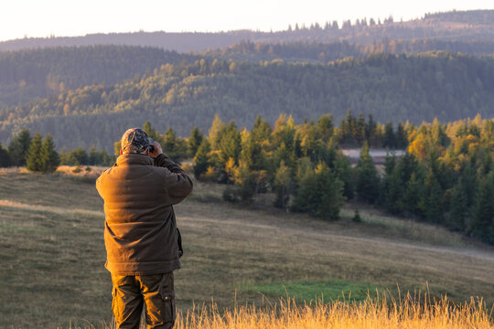 Man in camouflage looking through binoculars over mountain landscape at sunrise in autumn