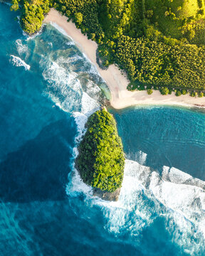 Aerial view of Pantai Goa Cina featuring a lush green rocky islet surrounded by turquoise ocean waves crashing against the sandy beach in Sumbermanjing Wetan, East Java, Indonesia.