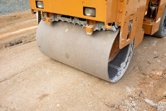 Vibratory roller flattens dirt pathway during municipal infrastructure works in daytime. Yellow compactor machine processes loose earth while maintaining consistent ground grade for pavement.
