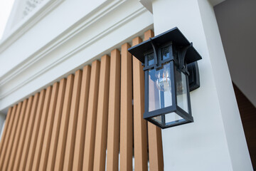 Naklejka premium Black vintage style wall lantern with glass panels and filament bulb mounted on white pillar of modern house, wooden slats behind, exterior lighting, outdoor decor, low angle view